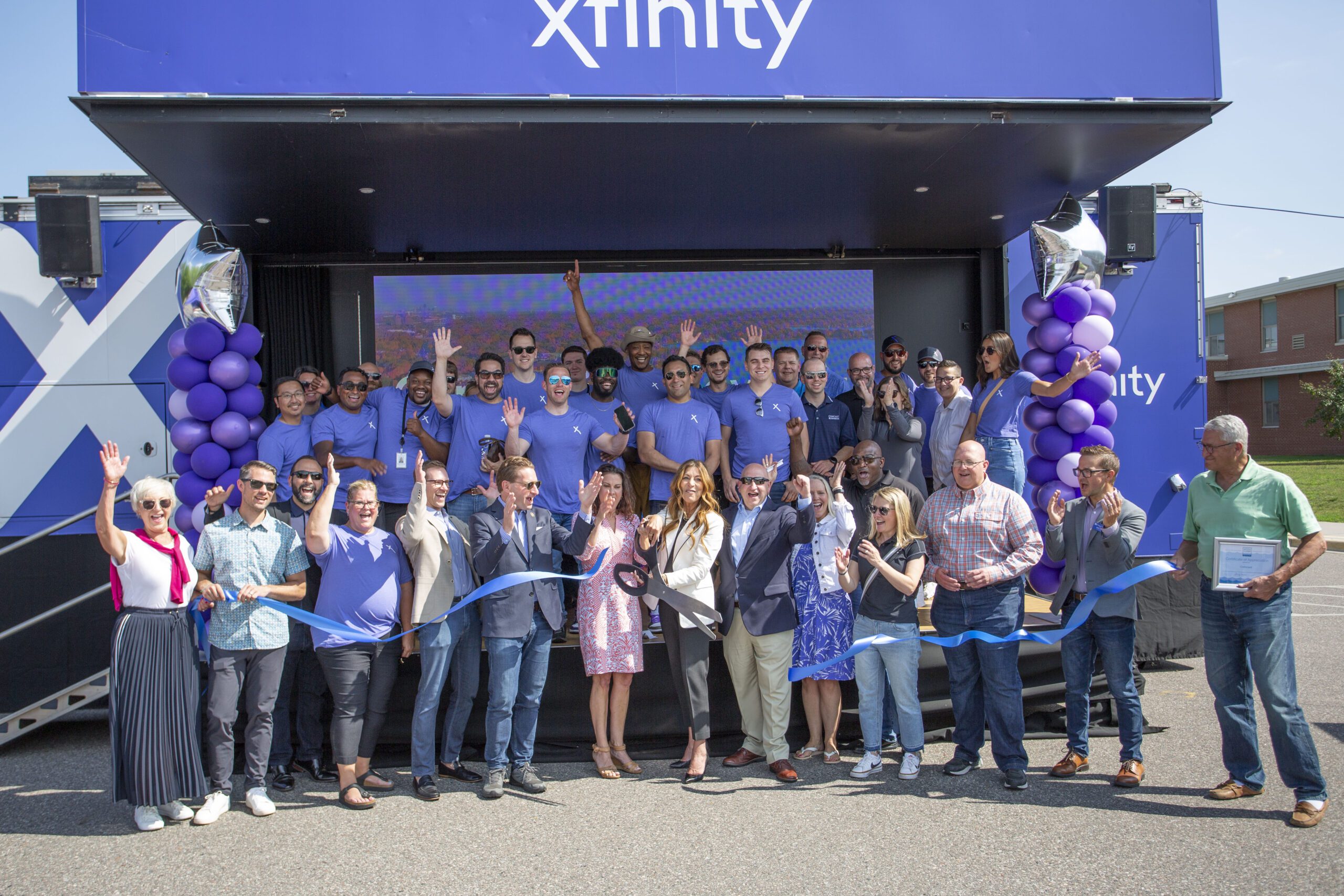 Large group of Comcast employees and city officials standing in front of a blue ribbon with hands up and smiles