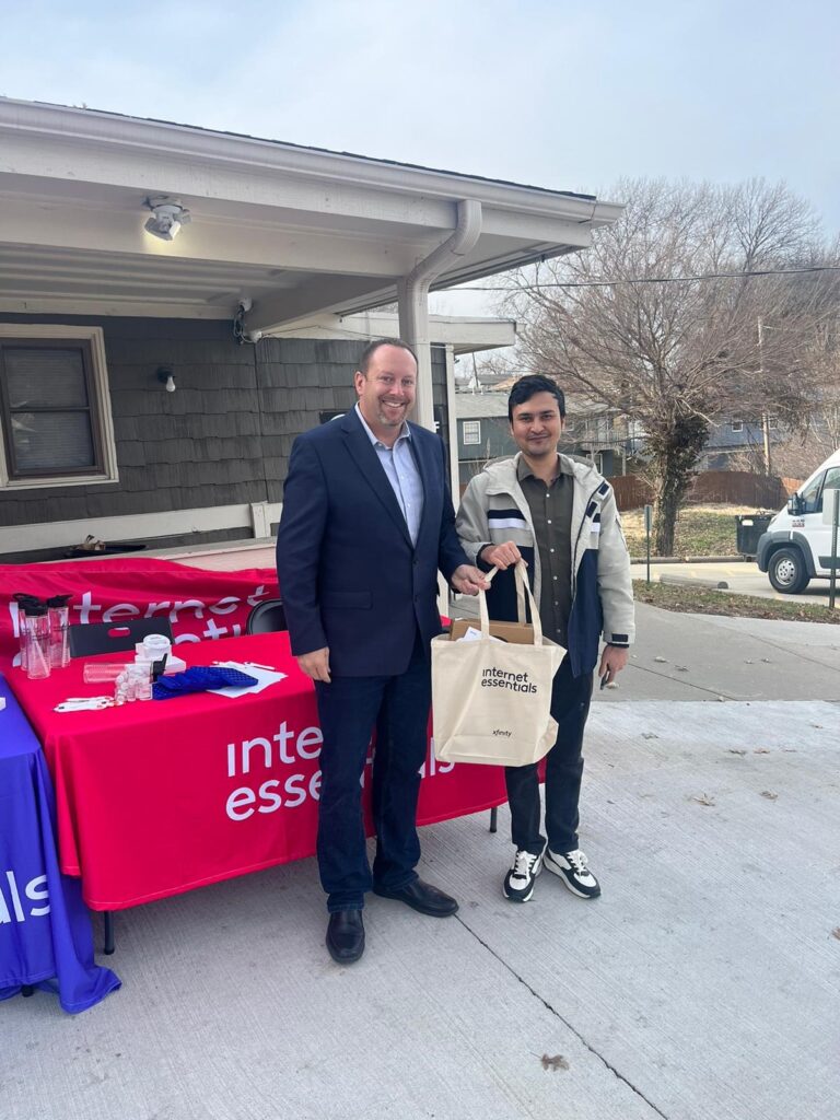 Jay and a recipient of our Internet Essentials program is standing in front of a red branded table, smiling for the camera and hold a branded Internet Essentials bag.