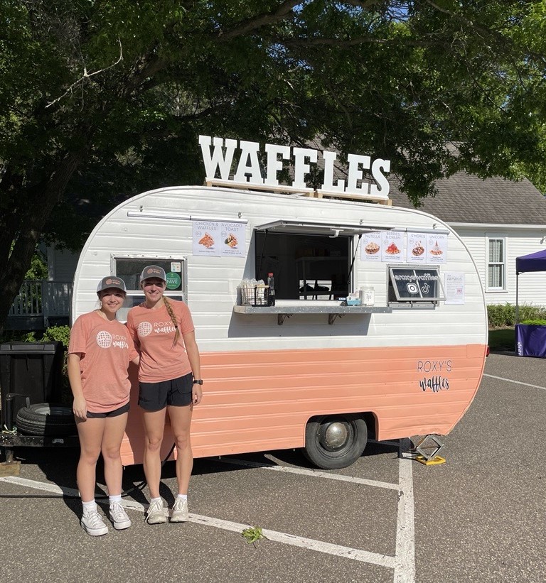 Co-owner and employee of Roxy's Waffles post in front of their peach-pink camper.