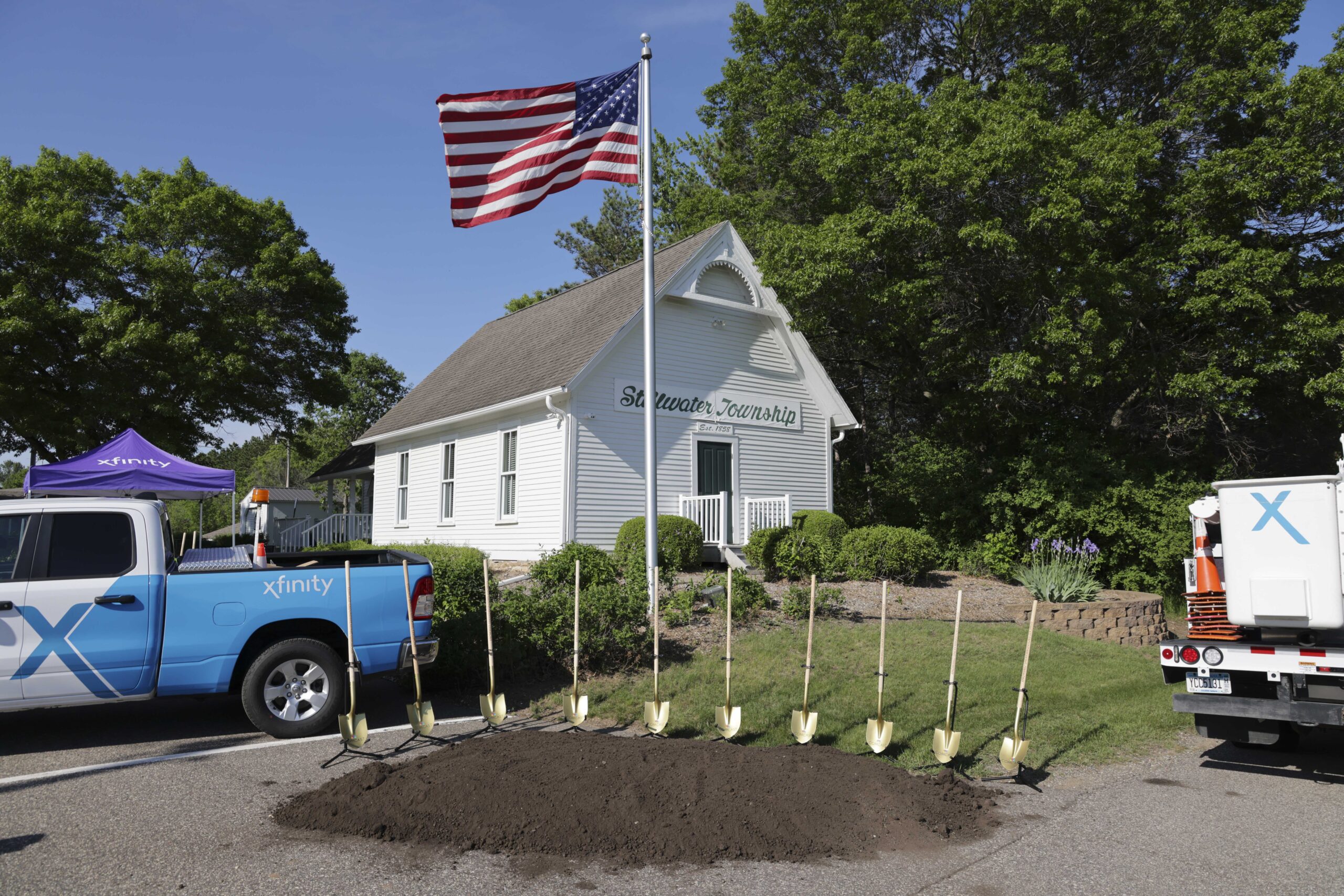 Xfinity branded vehicles parked in front of Stillwater Township's historica town hall. A large pile of dirt with golden shovel displayed on stands are set up between the two vehicles.